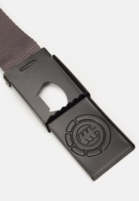 Correa de nailon gris con un regulador de plástico negro. El regulador cuenta con un logo en relieve y una sección recortada. Superficie texturizada con acentos suaves.