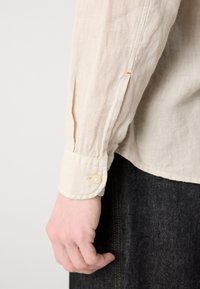 Gros plan d'une main portant un poignet de chemise beige à manches longues avec un bouton, reposant sur le côté au-dessus d'un pantalon texturé foncé.