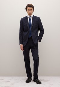 Dark navy suit, tailored with two buttons, paired with a white shirt and blue striped tie. Shoes are black leather with a smooth finish.