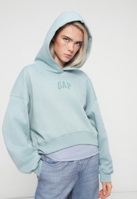 Felpa azzurra chiara con tasca frontale, logo GAP. Vestibilità oversize, polsini a costine, tessuto testurizzato, indossata sopra una maglietta a righe.