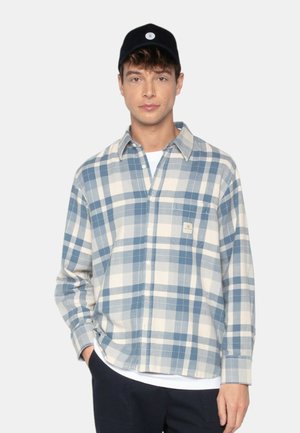 Un jeune homme portant une casquette noire, une chemise à carreaux bleus et blancs sur un T-shirt blanc, avec une main dans sa poche contre un fond uni.