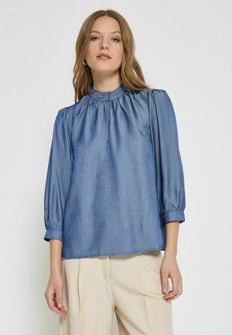 Denimbluse mit hohem Kragen, Raffung und Dreiviertelärmeln. Weiche Textur in einem mittleren Blauton, Knopfverschluss vorne.