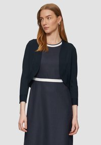 Cardigan cropped blu navy con maniche a tre quarti, polsini a costine e una texture liscia, abbinato a un vestito a pois con una cintura bianca.