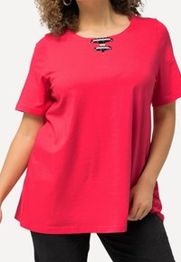 Camiseta roja de manga corta con escote en pico, que presenta un detalle decorativo de lazos en la parte delantera y un corte relajado. Textura suave.