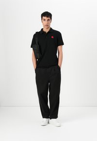 Μαύρο πόλο μπλουζάκι με κόκκινο λογότυπο, συνδυασμένο με χαλαρούς μαύρους παντελόνες και λευκά αθλητικά παπούτσια. Μοντέλο που κρατάει μια μικρή μαύρη τσάντα χιαστί.