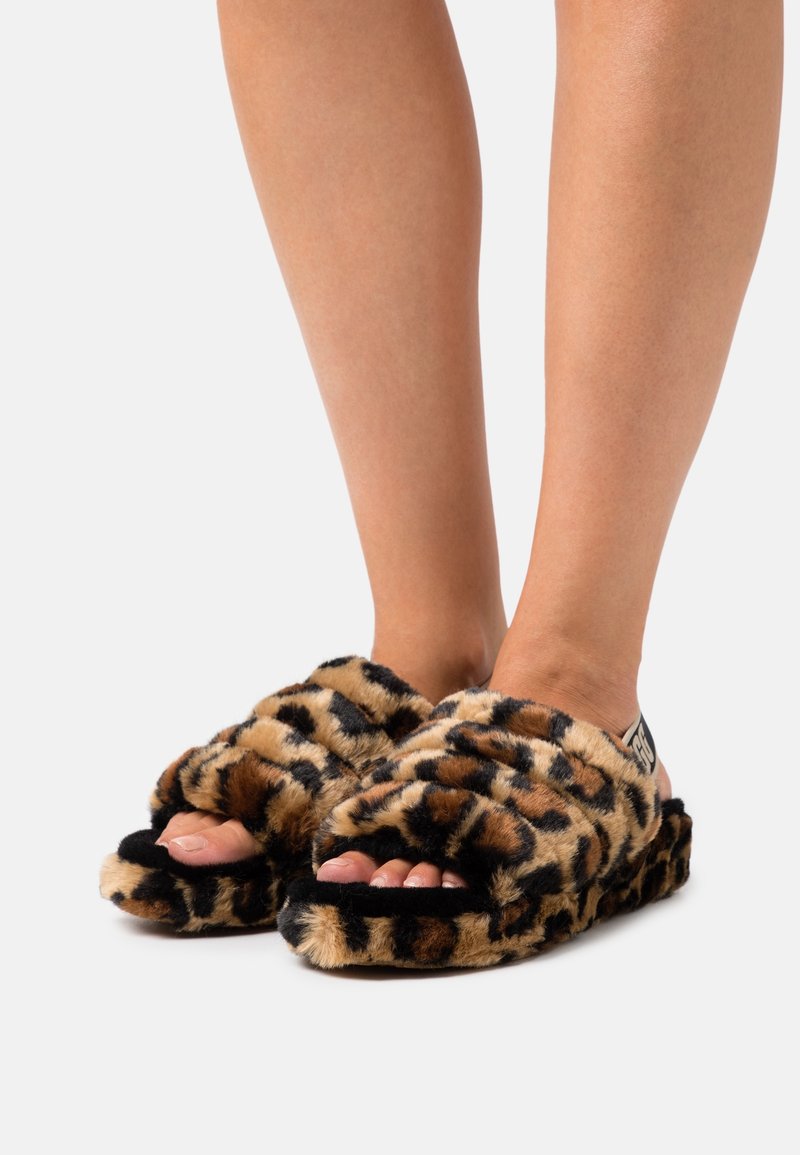 Jambes portant des pantoufles ouvertes douces avec un motif léopard marron et noir sur un fond uni.