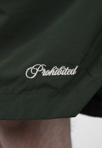 Tissu vert foncé avec broderie cursive blanche indiquant "Prohibital" près d'une couture cousue.