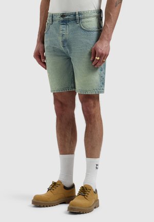 Man wearing light blue denim shorts, white socks, tan lace-up shoes, and a white shirt, standing against a plain background.