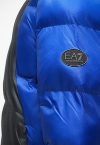 Zils pūkains mētelis ar teksturētu apdari, ar melnu sānu paneli un ovālu etiķeti, uz kuras ir norādīts "EA7 Emporio Armani."