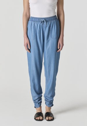 Mujer con pantalones jogger de cintura elástica color azul claro con bolsillos delanteros, combinados con sandalias negras sin talón, de pie frente a un fondo liso.