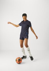 PUMA sötétkék sportfelszerelés, amely rövid ujjú felsőből és sortból áll, fekete focicipővel és fehér térd fölé érő zoknival párosítva, egy narancssárga focilabdával.