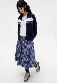 Μπλε μπουφάν με φερμουάρ και λευκές ρίγες, λευκό κοντό τοπ και μπλε καρό πλισέ φούστα. Φορεμένα με γκρι αθλητικά παπούτσια και μαύρες κάλτσες.