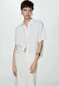 White linen button-up shirt with short sleeves and a relaxed fit. Features a row of gold buttons and is paired with a light-colored skirt.