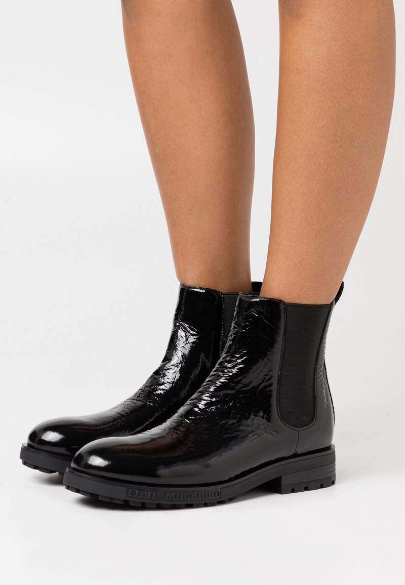 Bottines noires brillantes avec panneaux élastiques latéraux portées sur des jambes nues, avec des talons blocs bas et "Love Moschino" embossé sur les semelles.
