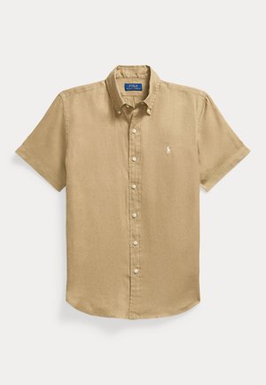 Man draagt een beige kortemouwen overhemd over een wit gebreid topje, witte broek met trekkoord, witte slip-on schoenen, houdt een bruine leren tas vast.