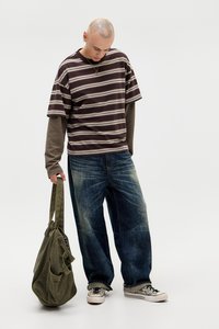 T-shirt de manga curta riscada em castanho e bege, sobreposta a uma camisa de manga longa. Jeans azul largos, sapatilhas prateadas e uma mala verde-azeitona.