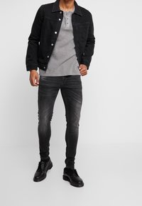 Chaqueta de mezclilla negra, camiseta henley gris y jeans skinny negros deslavados. Unos zapatos negros con cordones completan el atuendo. Diseño simple y casual.