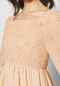 Žena v broskvově zbarvených šatech s malým oranžovým květinovým potiskem, s nabíraným živůtkem, čtvercovým výstřihem a nafouknutými rukávy do loktů.