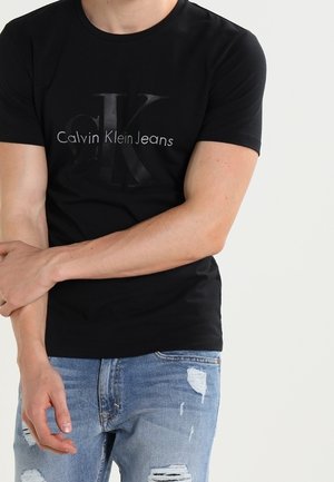 Schwarzes Baumwoll-T-Shirt mit großem grauem "CK"-Logo und dem Schriftzug "Calvin Klein Jeans" auf der Vorderseite. Kombiniert mit verwaschenen blauen Jeans.