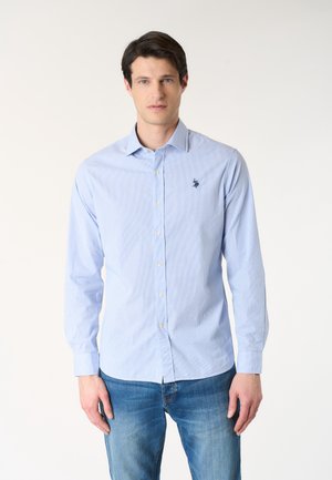 Uomo che indossa una camicia azzurro chiaro a maniche lunghe con bottoni, con un emblema scuro sul petto e jeans blu, in piedi davanti a uno sfondo semplice.