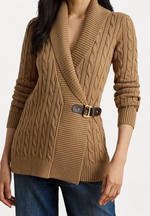 Kvinde iført en kamel-farvet kabelstrikket cardigan med en bred ribbet sjalskrave og en sidelæderbæltespænde, kombineret med blå jeans.