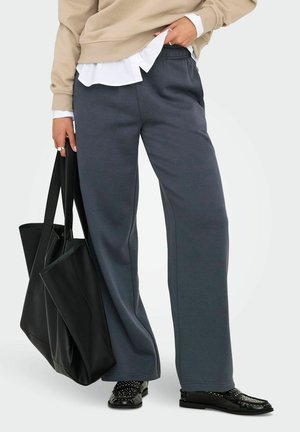 Grå brede sweatpants med elastisk talje, kombineret med en beige sweatshirt og en sort tote taske. Sorte sko med nitte-accenter.