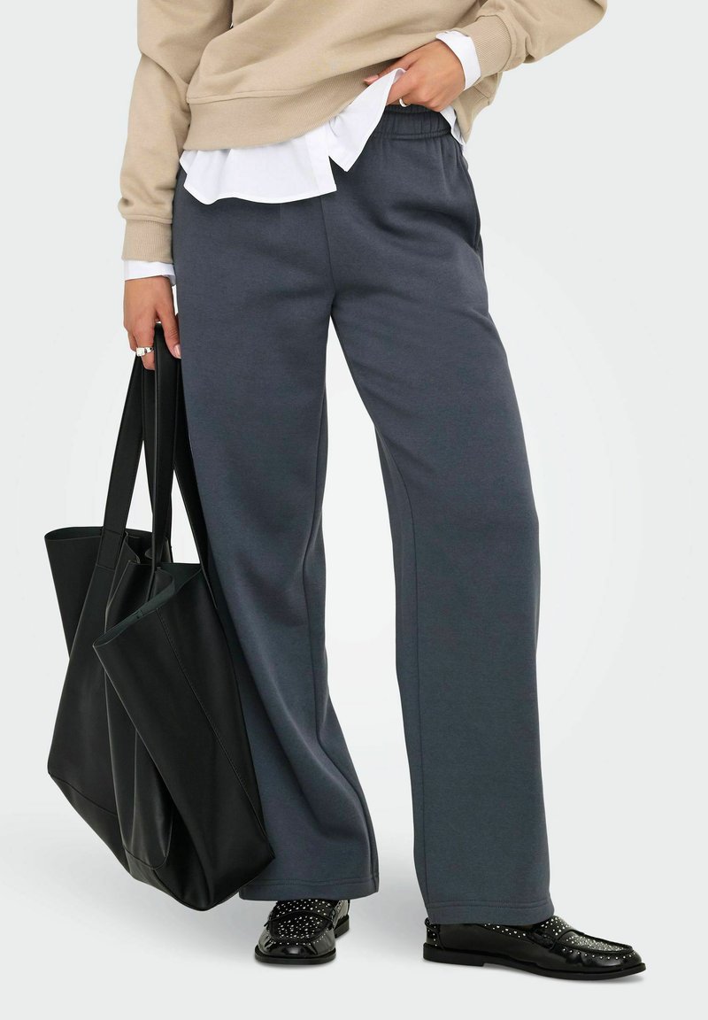 Grå brede sweatpants med elastisk talje, kombineret med en beige sweatshirt og en sort tote taske. Sorte sko med nitte-accenter.