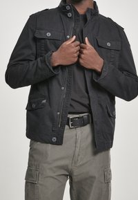 Svart jacka med krage och flera frontfickor, knapplist och dragkedja framtill, bärs över en mörk skjorta och matchas med cargobyxor.