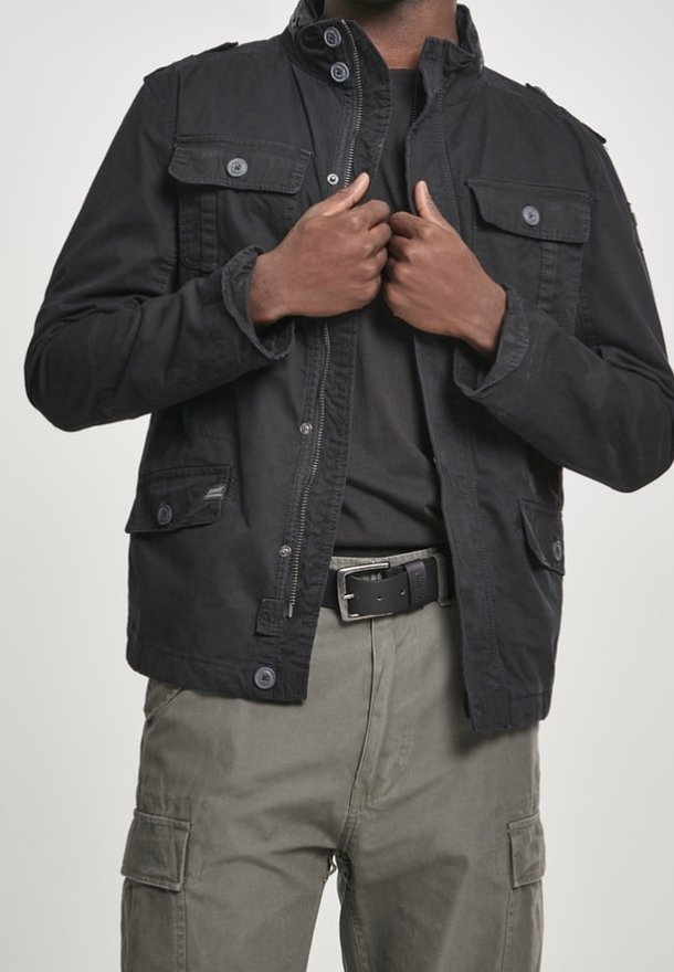 Svart jacka med krage och flera frontfickor, knapplist och dragkedja framtill, bärs över en mörk skjorta och matchas med cargobyxor.