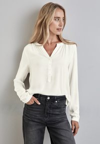 Chemise blanche à manches longues avec un col en V et cinq boutons, associée à un jean en denim foncé taille haute. Texture lisse, coupe décontractée.