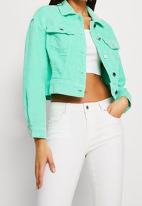 Miętowa kurtka z kordury, krótka, z dwoma kieszeniami na piersi, srebrnymi guzikami i teksturowym wykończeniem, zestawiona z białymi dopasowanymi spodniami.