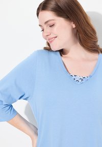 Lichtblauw, langärms top met een V-hals en knoopaccenten. Beschikt over een intricaat ontwerp onder de halslijn. Zacht, gestructureerd materiaal.