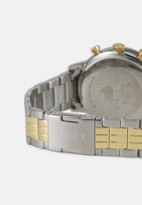 Timex laikrodis su nerūdijančio plieno juosta, apdailinta auksu, sidabriniu apvaliu korpusu ir matomais karūnėle bei mygtukais.