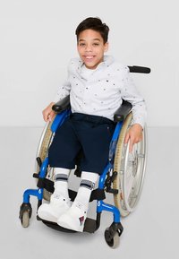 Dítě v modrém vozíku, oblečené v bílé košili s barevnými tečkami, tmavě modrých kalhotách a bílých botách s černými akcenty a pruhovanými ponožkami.