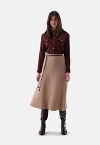 Woman with long brown hair wearing a burgundy button-up shirt, beige A-line skirt with a thin belt, and dark brown knee-high boots.