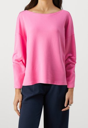 Kobieta ubrana w luźną, jasnoróżową bluzkę z długim rękawem, w połączeniu z ciemnogranatowymi spodniami, widoczna od ramion do bioder.