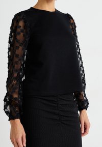 Svart topp med helfärgad överdel och transparenta ärmar med svarta blomsterapplikationer. Ärmsluten är lätt samlade. Texturerad svart kjol syns nedanför.