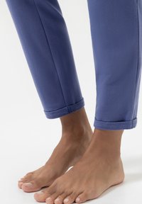 Strakke blauwe broek van zachte stof, met opgerolde boorden en een gladde textuur, gecombineerd met blote voeten zichtbaar onder de zoom.