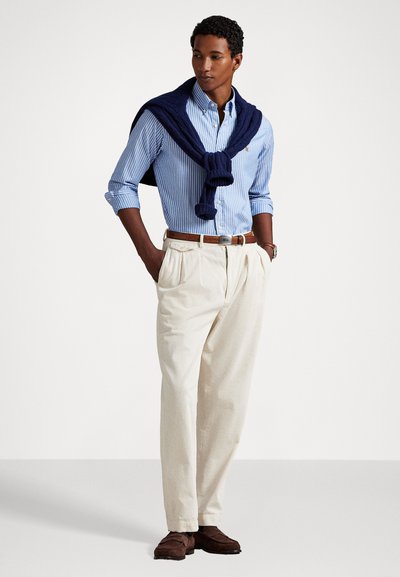 Chemise rayée bleu clair et blanche, pantalon plissé crème, pull marine drapé sur les épaules, ceinture marron et chaussures marron.