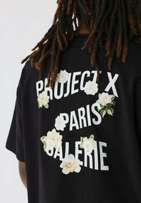 Zwarte katoenen t-shirt met witte tekst "PROJECT X PARIS GALERIE" en bloemenaccenten in crème en groen. Korte mouwen en een relaxte pasvorm.