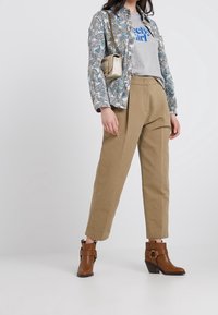 Mujer con una chaqueta de patrón azul claro y blanco sobre una camiseta gris, pantalones beige y botas tobilleras marrones, llevando un bolso pequeño.