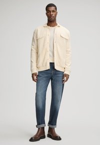 Cardigan en tricot crème avec des motifs en câble, fermeture à boutons, associé à un jean en denim bleu clair et des bottines en cuir marron.