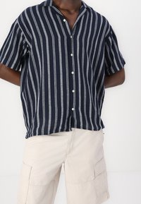 Donkerblauw shirt met korte mouwen en witte verticale strepen, knoopsluiting aan de voorkant en zacht, lichtgewicht materiaal. Losse pasvorm met een kraag.