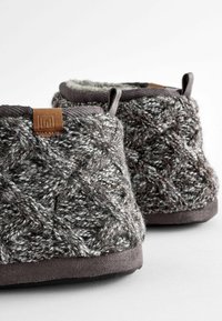 Gestrickte graue und weiße Hausschuhe mit einem strukturierten Muster, gepolstertem Innenfutter, braunem Lederakzent und einer glatten dunklen Sohle.