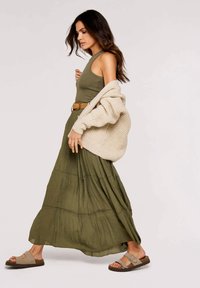 Vestido maxi verde oliva con diseño de volantes y un suéter de punto beige texturizado caído sobre un hombro. Combinado con sandalias marrón claro.