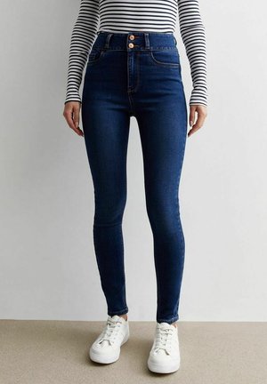 Kobieta w ciemnoniebieskich, dopasowanych jeansach z wysokim stanem, w połączeniu z białymi trampkami i czarno-białą, paskowaną bluzką z długim rękawem.