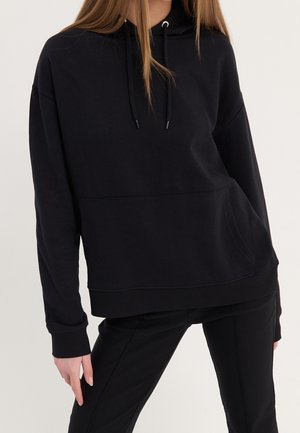 Femme portant un sweat à capuche noir avec une poche avant et un pantalon noir, debout devant un fond clair uni.