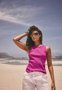 Priliehavý vrúbkovaný top v jasnej ružovej farbe, v kombinácii s bielymi šortkami. Modelka má na sebe oversized slnečné okuliare a pózuje proti plážovému pozadiu.