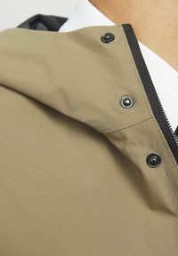 Khaki jack met capuchon, met een gladde textuur, drukknoopsluitingen en zwarte accenten langs de rits. Close-up weergave focust op de details van de halslijn.