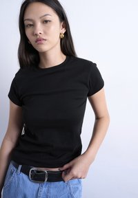 Νεαρή γυναίκα με μακριά σκούρα μαλλιά φοράει μαύρο μπλουζάκι, χρυσά σκουλαρίκια κρίκους, ανοιχτό μπλε τζιν και μαύρη ζώνη, στέκεται μπροστά σε λευκό φόντο.